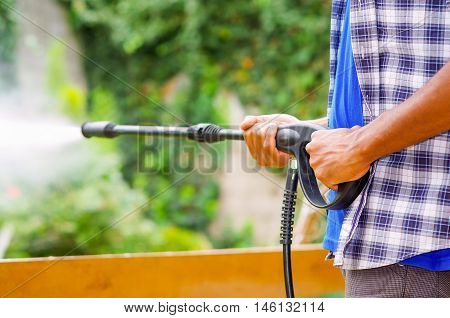 Closeup arms of man wearing square pattern blue and white shirt holding high pressure water gun, pointing towards green garden.