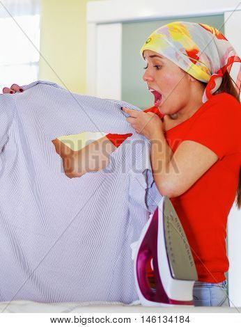 Young charming woman wearing colorful headscarf holding up office shirt with hole bruned through fabric, shocked facial expression, laundry housework concept.