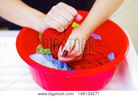 Closeup womans hands handwashing clothes in red plastic washbucket, scrubbing and squeezing fabrics, laundry housework concept.