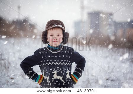 Winter portrait of little kid boy wearing a knitted sweater with deers outdoors during snowfall. Active outdoors leisure with children in winter on cold snowy days
