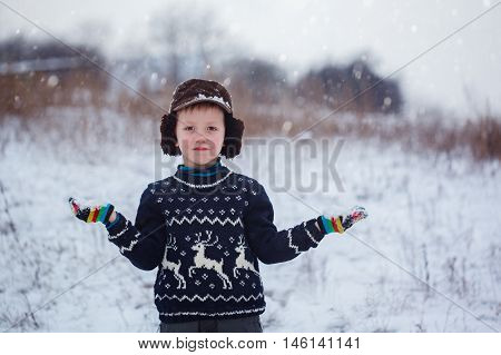 Winter portrait of little kid boy wearing a knitted sweater with deers outdoors during snowfall. Active outdoors leisure with children in winter on cold snowy days.