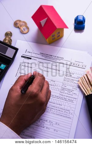 Indian man or accountant person filing Indian income tax returns form or ITR document showing indian currency, house model, toy car and calculator over white table top, selective focus
