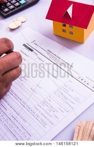 Indian man or accountant person filing Indian income tax returns form or ITR document showing indian currency, house model, toy car and calculator over white table top, selective focus