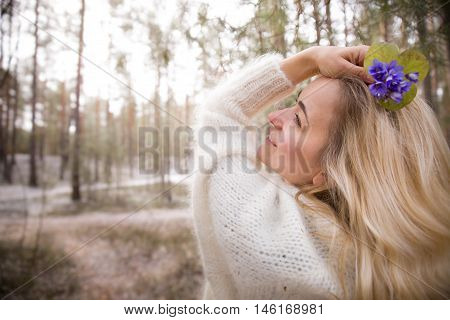 Young blond woman in white warm cardigan standing near an old tree and holding beautiful snowdrops in hands. First spring flowers in forest. Beginning of spring in a forest. Sunlight in a pine forest