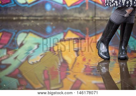 Girl in rubber boots standing in the puddle in the street. Woman in grey rubber boots splashing in a puddle after rain. Pair of grey rubber boots in a big puddle with graffiti reflections.