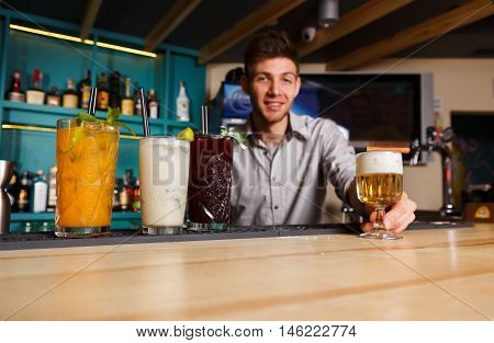 Young handsome Barman offers exotic sweet non-alcoholic cocktails in restaurant. Professional male bartender at work in bar made refreshing drinks. Selective focus on glass of cream soda