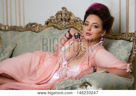 beautiful brunette with curvaceous Rococo hair rings and beads lies on a beautiful sofa in pink peignoir