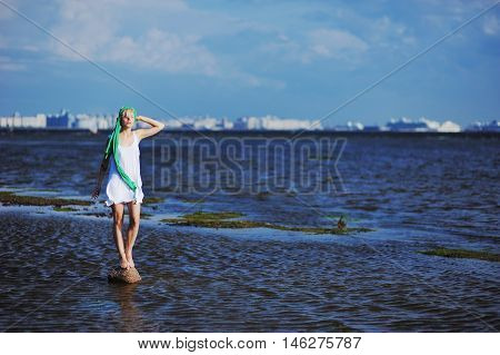 blonde in a white dress with a green scarf is standing on a stone in the middle of the water