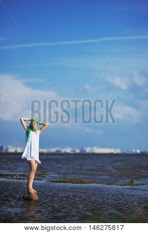 blonde in a white dress with a green scarf is standing on a stone in the middle of the water