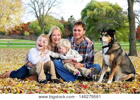 A happy family of four people and their pet dog are posing for a portrait in the fallen leaves in the park as their children goof around.