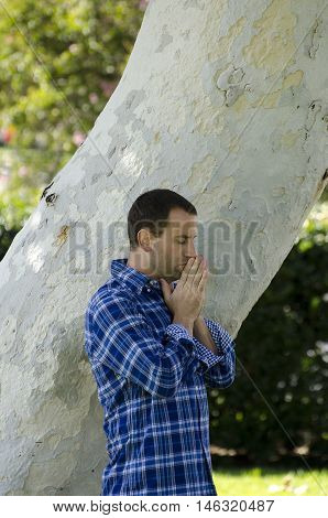 Young man praying in a park by a large tree trunk.