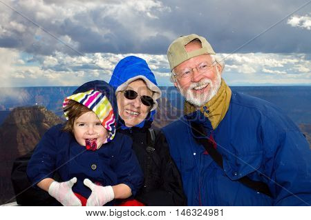 Happy grandparents hold their young granddaughter at the overlook of Bright Angel at the North Rim of the Grand Canyon. All three are dressed in winter clothes and snow is falling around them.