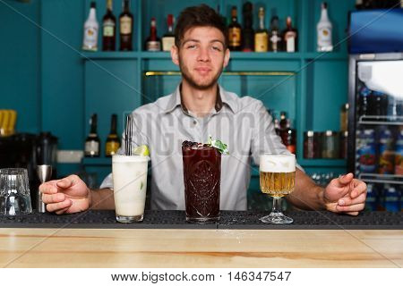 Young handsome Barman offers exotic sweet non-alcoholic cocktails in restaurant. Professional bartender at work in bar made refreshing drinks. Selective focus on glasses of cream soda and berry mix