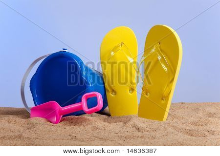 ถังสีฟ้า สีชมพูพลั่ว และ flipflops สีเหลืองบนหาดทรายกับพื้นสีน้ำเงิน