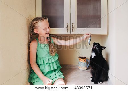 Child girl feeding a black cat in the kitchen. Black cat eating fish.