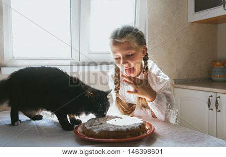 Child girl feeding a black cat in the kitchen.