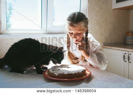 Child girl feeding a black cat in the kitchen.