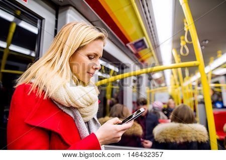 Beautiful young blond woman in red coat in subway train, holding a smart phone, texting
