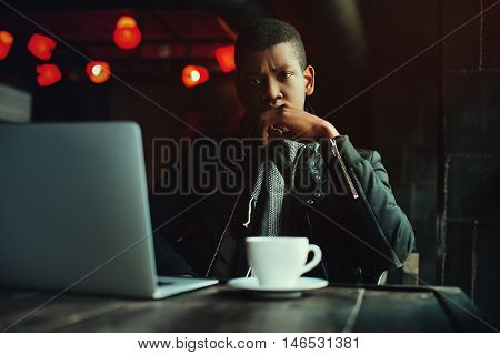 Indoor portrait of young black man sitting in cafe, drinking coffee or tea and working with lap top. Model looking at camera. Toned.