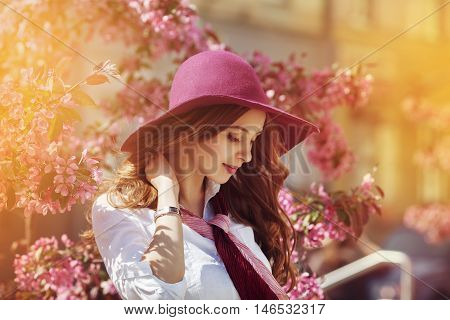 Outdoor portrait of young beautiful happy smiling lady posing near flowering tree. Model wearing stylish accessories and clothes. Girl closed her eyes. Female beauty and fashion concept. City lifestyle