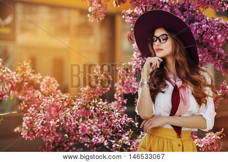 Outdoor portrait of young beautiful fashionable lady posing near flowering tree. Model wearing stylish accessories and clothes. Girl closed her eyes. Female beauty, fashion. City lifestyle. Copy space.