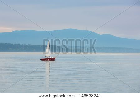 Red sailboat sailing in calm blue bay in Bellingham, Washington during purple sunset with mountain