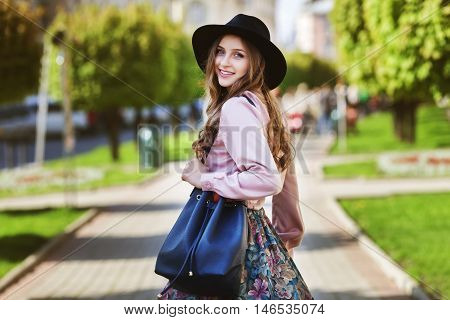 Outdoor portrait of young beautiful fashionable happy smiling lady walking on street of the old city. Model wearing stylish fedora hat and clothes. Girl looking at camera. Female fashion. City lifestyle