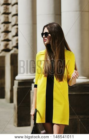 Outdoor portrait of young beautiful lady walking on the street. Model wearing sunglasses and stylish yellow summer dress. Girl looking aside. Female fashion concept. City lifestyle. Sunny day. Waist up.