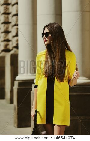 Outdoor portrait of young beautiful lady walking on the street. Model wearing sunglasses and stylish yellow summer dress. Girl looking aside. Female fashion. City lifestyle. Sunny day. Waist up. Toned.