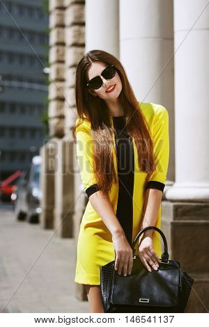 Outdoor portrait of young beautiful happy lady walking on street. Model wearing sunglasses and stylish yellow summer dress. Girl looking at camera. Female fashion. City lifestyle. Sunny day. Waist up.