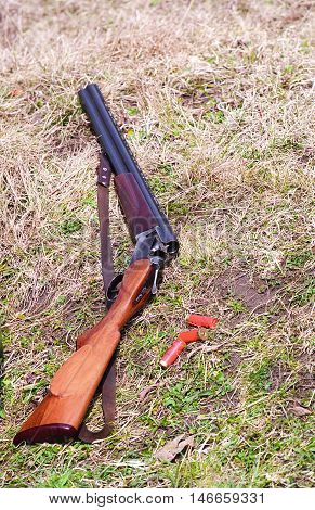 A view of a double barrel shotgun lying on the ground with several red shotgun shells nearby. The shotgun is open ready to be loaded.