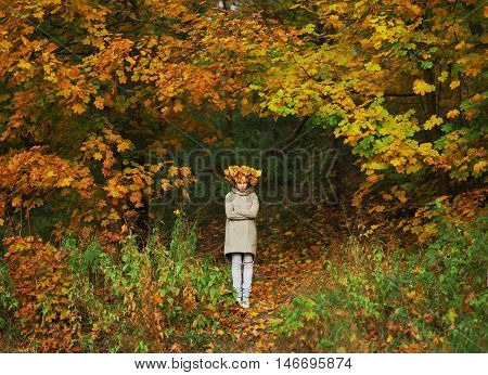 Young frowning girl with a wreath of autumn maple leaves on the head is on the forest path between trees