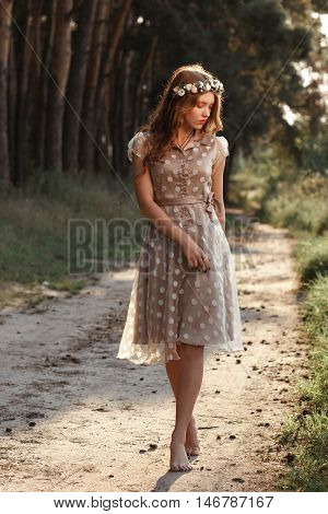 Young woman in wreath walking in forest barefoot. Beautiful girl in polka-dot dress relaxing in nature, she steps on path with pine cones, free space.
