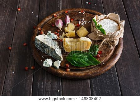 Cheese delikatessen closeup on rustic wood. Wooden round desk with blue cheese roquefort, parmesan, camembert and brie cuts decorated with garlic, pomegranate and basil, french cuisine ingredients
