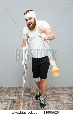 Mad bandaged young man standing and pointing on you with crutches over white background