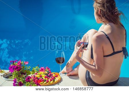 Pretty woman in swimsuit with glass of red wine fruit platter sitting back relaxing tanning and dreaming near pool with blue water in summer