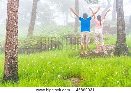 Young couple of love spead hand and relaxed in the midst of pink flower field with green meadaw and moss rock in pine forest that was covered by morning fog. Bolaven Plateau Southern fo Laos.