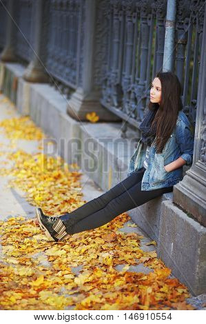 Young long-haired brunette sitting on a concrete curb around the cast-iron fence, under the feet of yellow autumn maple leaves.