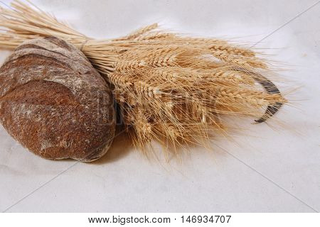 bunch of mown wheat ears with vintage handmade reaper hook sickle and rye dark french fresh bread loaf on white linen tablecloth