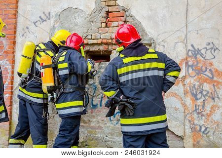 View From Behind On Firefighter In Uniform With Full Safety Gear, Using Crowbar To Break Brick Wall.
