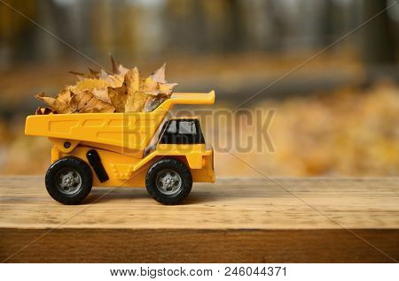 A Small Toy Yellow Truck Is Loaded With Yellow Fallen Leaves. The Car Stands On A Wooden Surface Aga