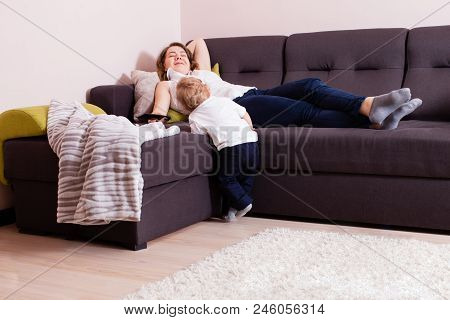 Happy Mother Laying On Back On Sofa With Her Baby Son Standing Near Her. Young Mother Laying On The 