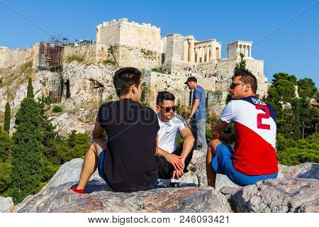 Boys Seated On A Rock And Chatting Against The Acropoilis Ruins In Athens, Greece On A Summer Aftern