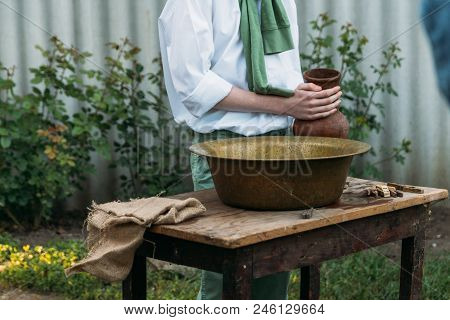 A Boy In A White Shirt And A Green Sweater, Holds A Clay Jug, Pours Water Out Of It Into A Copper Ba