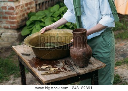 A Boy In A White Shirt And A Green Sweater, Holds A Clay Jug, Pours Water Out Of It Into A Copper Ba