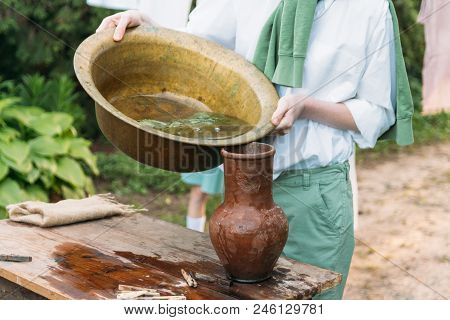 A Boy In A White Shirt And A Green Sweater, Holds A Clay Jug, Pours Water Out Of It Into A Copper Ba