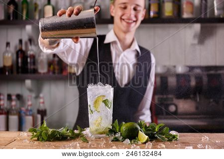A Smiling Barman Pours Water Into A Glass Of Refreshing Mojito, Decorated With Mint And Limes.