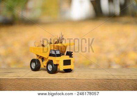 A Small Toy Yellow Truck Is Loaded With Yellow Fallen Leaves. The Car Stands On A Wooden Surface Aga