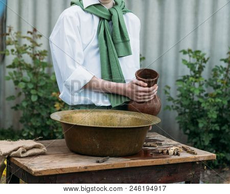 A Boy In A White Shirt And A Green Sweater, Holds A Clay Jug, Pours Water Out Of It Into A Copper Ba