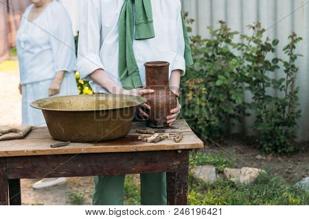 A Boy In A White Shirt And A Green Sweater, Holds A Clay Jug, Pours Water Out Of It Into A Copper Ba